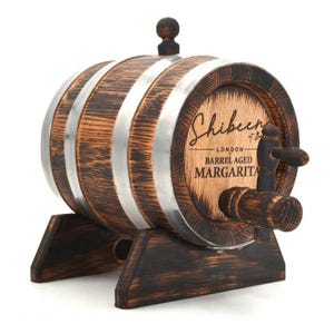 May include: A small, wooden barrel with metal bands and a spigot. The barrel is dark brown with a lighter wood front, featuring the text "Shibeen London Barrel Aged Margarita". It sits on a matching wooden stand.