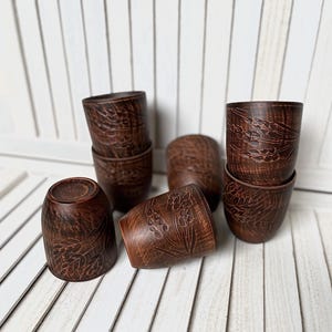 May include: A collection of handmade, dark brown ceramic cups. Each cup features a detailed floral pattern carved into the surface. The cups are arranged on a white slatted surface, showcasing their rustic charm and craftsmanship.