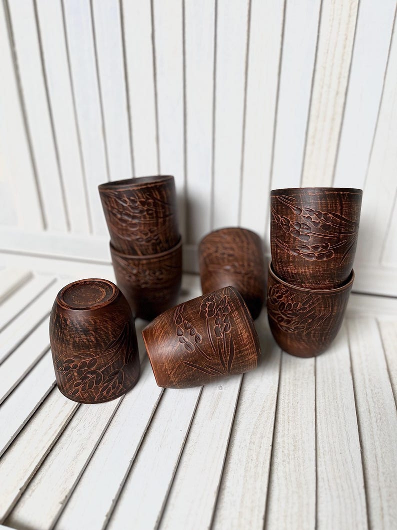 May include: A collection of handmade, dark brown ceramic cups. Each cup features a detailed floral pattern carved into the surface. The cups are arranged on a white slatted surface, showcasing their rustic charm and craftsmanship.