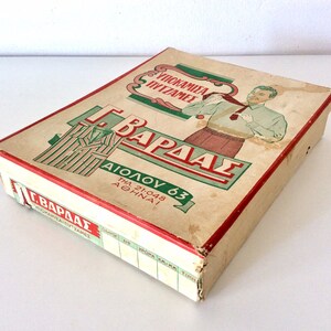 May include: Vintage rectangular box with a red trim, featuring Greek text and illustrations. The box has a worn, aged appearance, with a drawing of a man and a pajama set. The text includes the name "G. Vardas" and the address "Aiolou 63, Athinai".