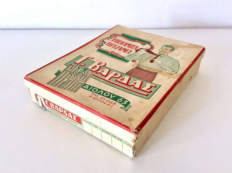 May include: Vintage rectangular box with a red trim, featuring Greek text and illustrations. The box has a worn, aged appearance, with a drawing of a man and a pajama set. The text includes the name "G. Vardas" and the address "Aiolou 63, Athinai".