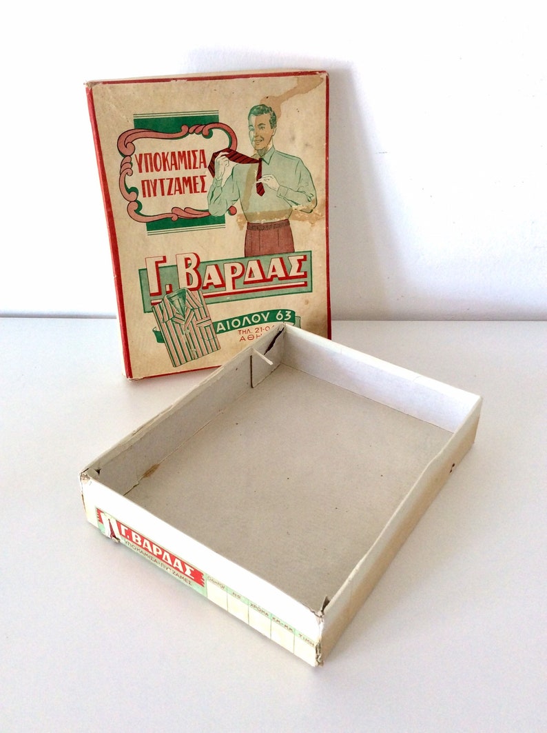 May include: Vintage cardboard box with a red and green label featuring a man adjusting his tie. The box is open, revealing its empty interior. The text on the box is in Greek.