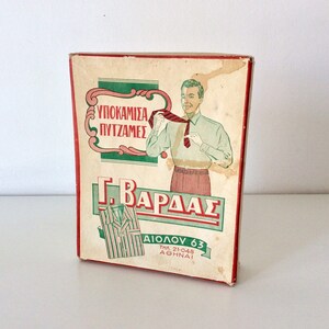 May include: Vintage box with a retro illustration of a man adjusting his tie. The box features Greek text and a green and red color scheme. The box is slightly worn, suggesting its age.
