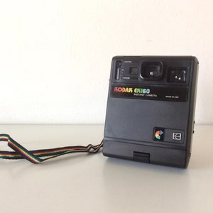 Peut inclure: Appareil photo instantané Kodak EK160 noir avec une dragonne rayée arc-en-ciel. L'appareil photo a un flash, un objectif et le logo Kodak aux couleurs de l'arc-en-ciel. Fabriqué aux États-Unis.