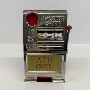 May include: Silver-colored metal toy bank shaped like a slot machine with a red lever. The front of the bank has a red button, three windows with colorful images, and the text "JACKPOT BANK". Below the windows is the text "1¢ 5¢ 10¢" and below that is a gold rectangle with the text "AID CORP."