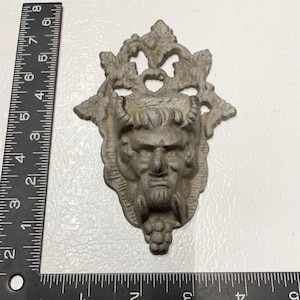 May include: A gray metal wall decor featuring a stylized face with horns and a grape cluster. The face is surrounded by a decorative design of leaves and vines.