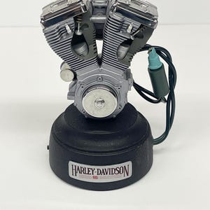 May include: A silver and chrome Harley-Davidson engine model. The model sits on a black base with the Harley-Davidson logo. A green and black wire with a blue tip extends from the engine.