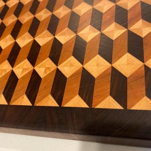 May include: A rectangular wooden cutting board featuring an intricate geometric pattern of three-dimensional cubes. The design is crafted from contrasting light, medium, and dark wood tones, framed by a solid dark wood border along the edges.
