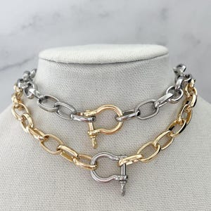 May include: Two chunky chain necklaces on a mannequin. One is gold-toned, the other silver-toned. Both have large oval links and a decorative clasp. The necklaces are likely fashion jewellery.