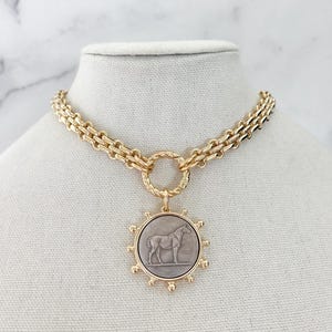 May include: A gold-toned chain necklace featuring a circular pendant with a grey centre inlay. The pendant displays a relief of a horse and is surrounded by small gold spheres. The chain consists of interlocking links connected to a twisted ring.