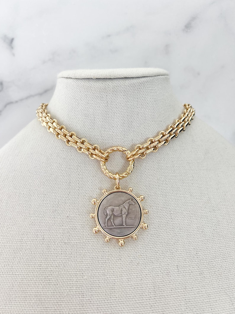 May include: A gold-toned chain necklace featuring a circular pendant with a grey centre inlay. The pendant displays a relief of a horse and is surrounded by small gold spheres. The chain consists of interlocking links connected to a twisted ring.