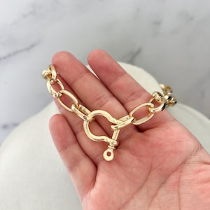 Puede incluir: Un collar de cadena dorado con un cierre grande. El collar est&aacute; hecho de eslabones gruesos de forma ovalada. El cierre tiene un dise&ntilde;o en forma de coraz&oacute;n con cierre de pasador. El collar se muestra sobre una superficie blanca.