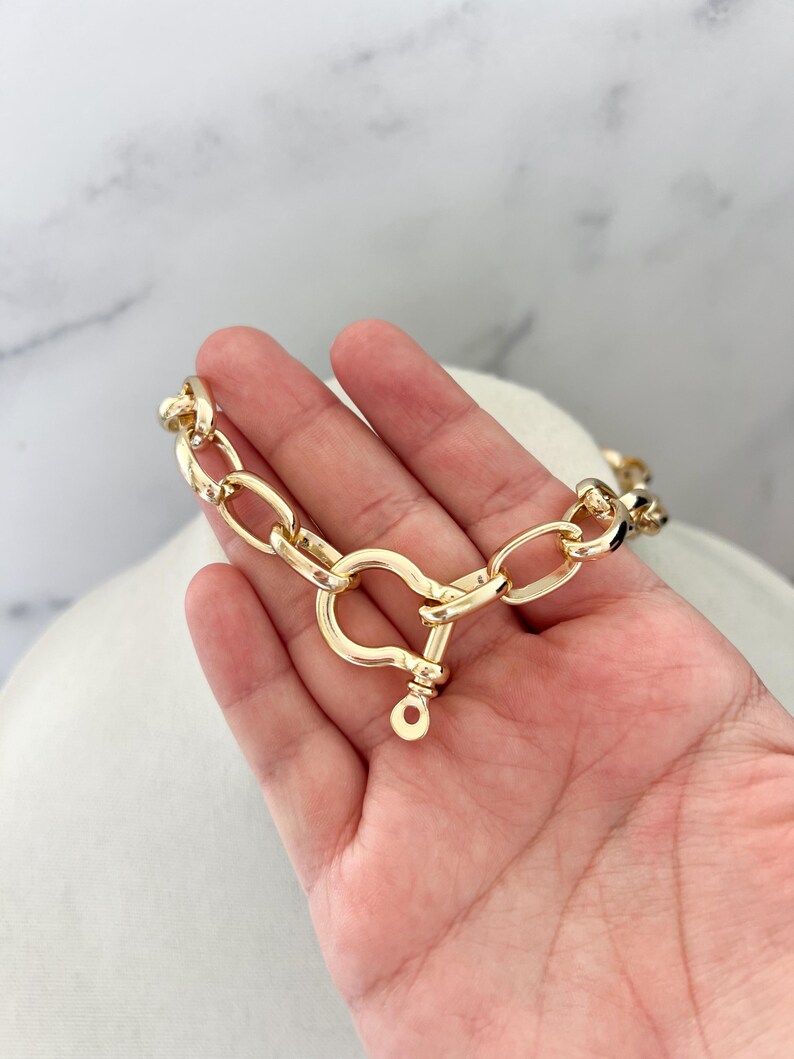 Puede incluir: Un collar de cadena dorado con un cierre grande. El collar est&aacute; hecho de eslabones gruesos de forma ovalada. El cierre tiene un dise&ntilde;o en forma de coraz&oacute;n con cierre de pasador. El collar se muestra sobre una superficie blanca.