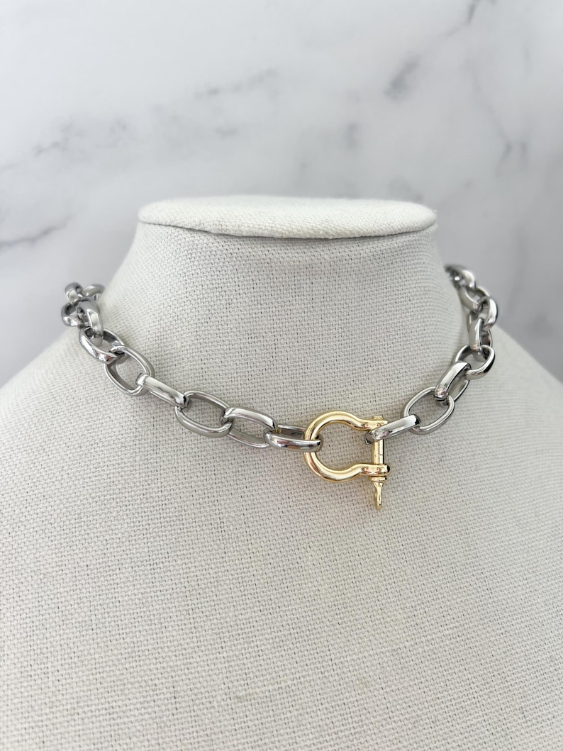 Puede incluir: Un collar de cadena plateada con un cierre dorado. El collar se exhibe sobre un busto de maniqu&iacute; blanco sobre un fondo de m&aacute;rmol. Los eslabones de la cadena son ovalados y gruesos, creando un aspecto audaz y moderno. Aproximadamente 40 cm de largo.