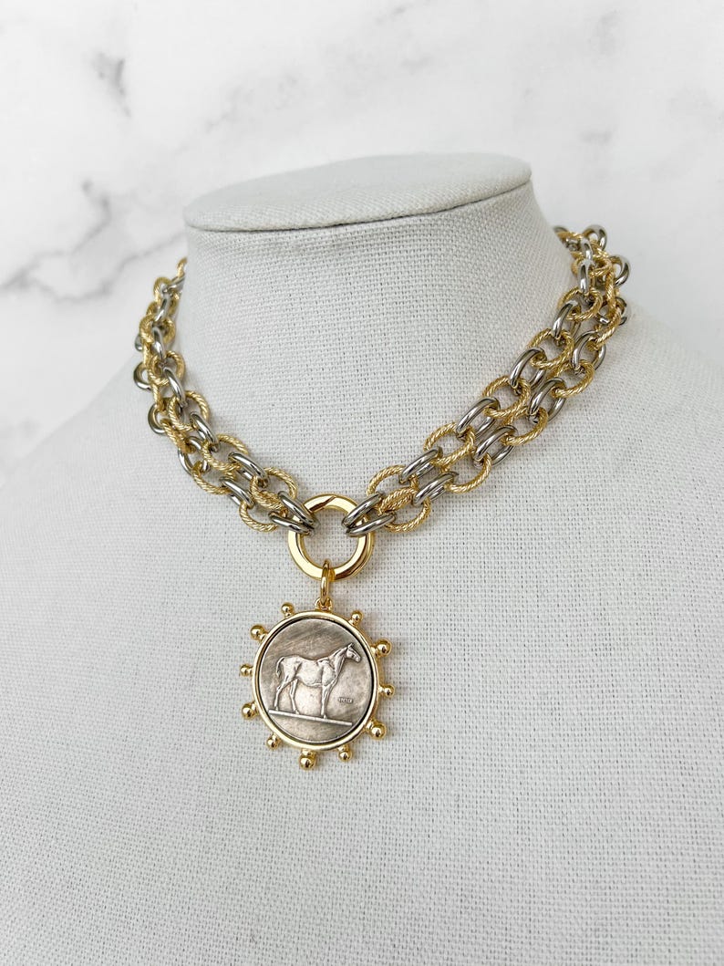 May include: A gold and silver chain necklace with a round pendant featuring a silver horse design. The pendant has a gold rim with small gold balls around the edge.
