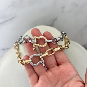Puede incluir: Dos pulseras de cadena en tonos dorados y plateados. Cada pulsera presenta un cierre grande y curvado. Las pulseras se exhiben sobre una superficie blanca, resaltando su dise&ntilde;o y acabado met&aacute;lico. La joyer&iacute;a es adecuada para el uso diario.