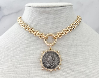 Puede incluir: Un collar de cadena en tono dorado con un colgante circular de centro gris oscuro. El colgante muestra un escudo en relieve rodeado por una corona y pequeñas perlas blancas en el borde exterior, suspendido de un eslabón circular dorado.