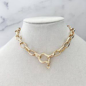 Puede incluir: Un collar de cadena de color dorado con un cierre de cierre grande. El collar se exhibe sobre un busto de maniqu&iacute; blanco contra un fondo de m&aacute;rmol blanco y gris. Los eslabones de la cadena tienen forma ovalada.