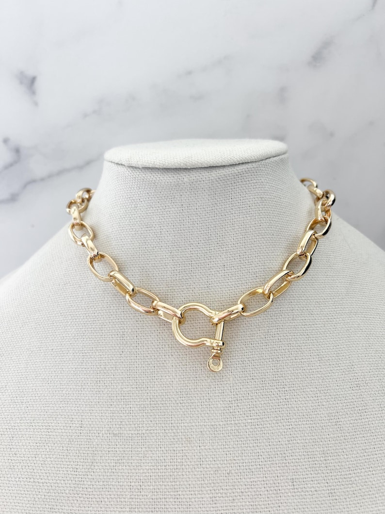Puede incluir: Un collar de cadena de color dorado con un cierre de cierre grande. El collar se exhibe sobre un busto de maniqu&iacute; blanco contra un fondo de m&aacute;rmol blanco y gris. Los eslabones de la cadena tienen forma ovalada.