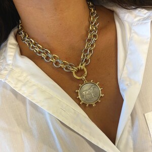May include: A gold and silver chain necklace with a large pendant featuring a silver horse on a gold coin-shaped disc. The pendant is surrounded by a gold circle with small gold beads.