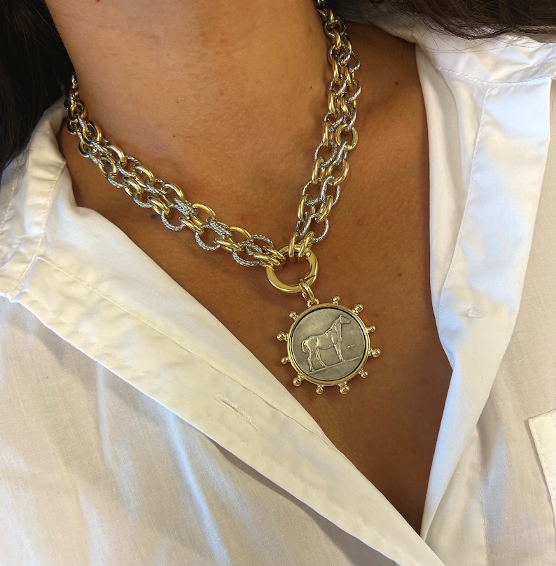 May include: A gold and silver chain necklace with a large pendant featuring a silver horse on a gold coin-shaped disc. The pendant is surrounded by a gold circle with small gold beads.