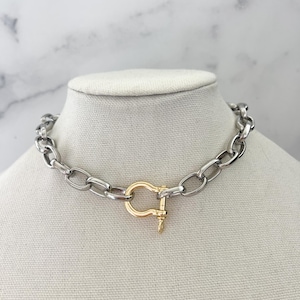 Puede incluir: Un collar de cadena plateada con un cierre dorado. El collar se exhibe sobre una cabeza de maniqu&iacute; blanca sobre un fondo de m&aacute;rmol blanco y gris. Los eslabones de la cadena son de forma ovalada.