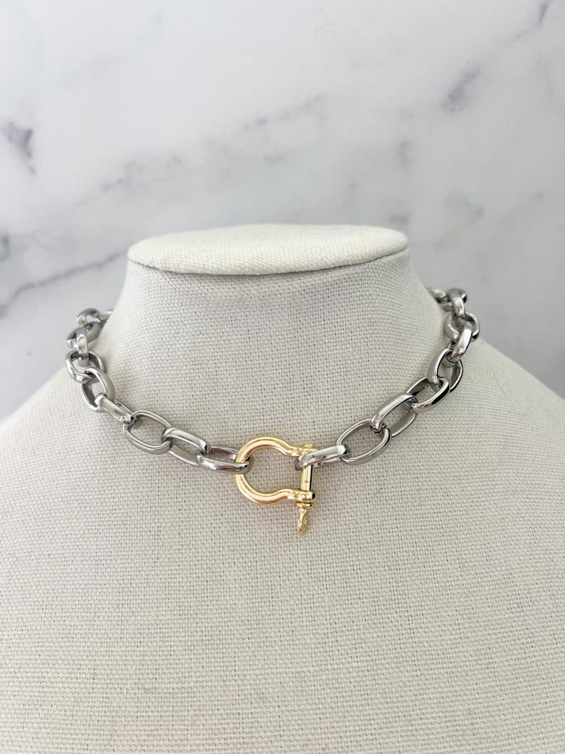 Puede incluir: Un collar de cadena plateada con un cierre dorado. El collar se exhibe sobre una cabeza de maniqu&iacute; blanca sobre un fondo de m&aacute;rmol blanco y gris. Los eslabones de la cadena son de forma ovalada.