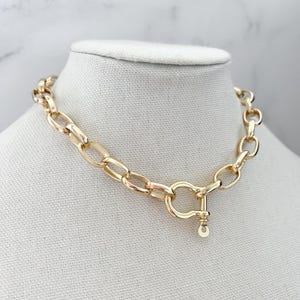 Puede incluir: Collar de cadena dorada con un dise&ntilde;o de eslabones grandes. El collar presenta un cierre de cierre &uacute;nico con un peque&ntilde;o dije redondo. La joya se exhibe sobre un busto de maniqu&iacute; blanco sobre un fondo de m&aacute;rmol.