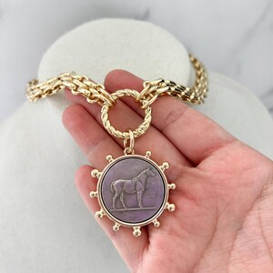 May include: A gold-toned chain necklace featuring a large, circular pendant with a purple centre. The pendant displays a relief of a horse standing on a horizontal line, surrounded by a sunburst-style border with small spheres at each point.