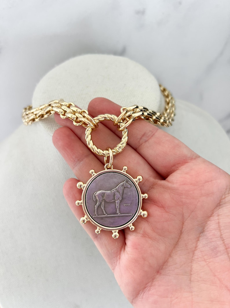 May include: A gold-toned chain necklace featuring a large, circular pendant with a purple centre. The pendant displays a relief of a horse standing on a horizontal line, surrounded by a sunburst-style border with small spheres at each point.