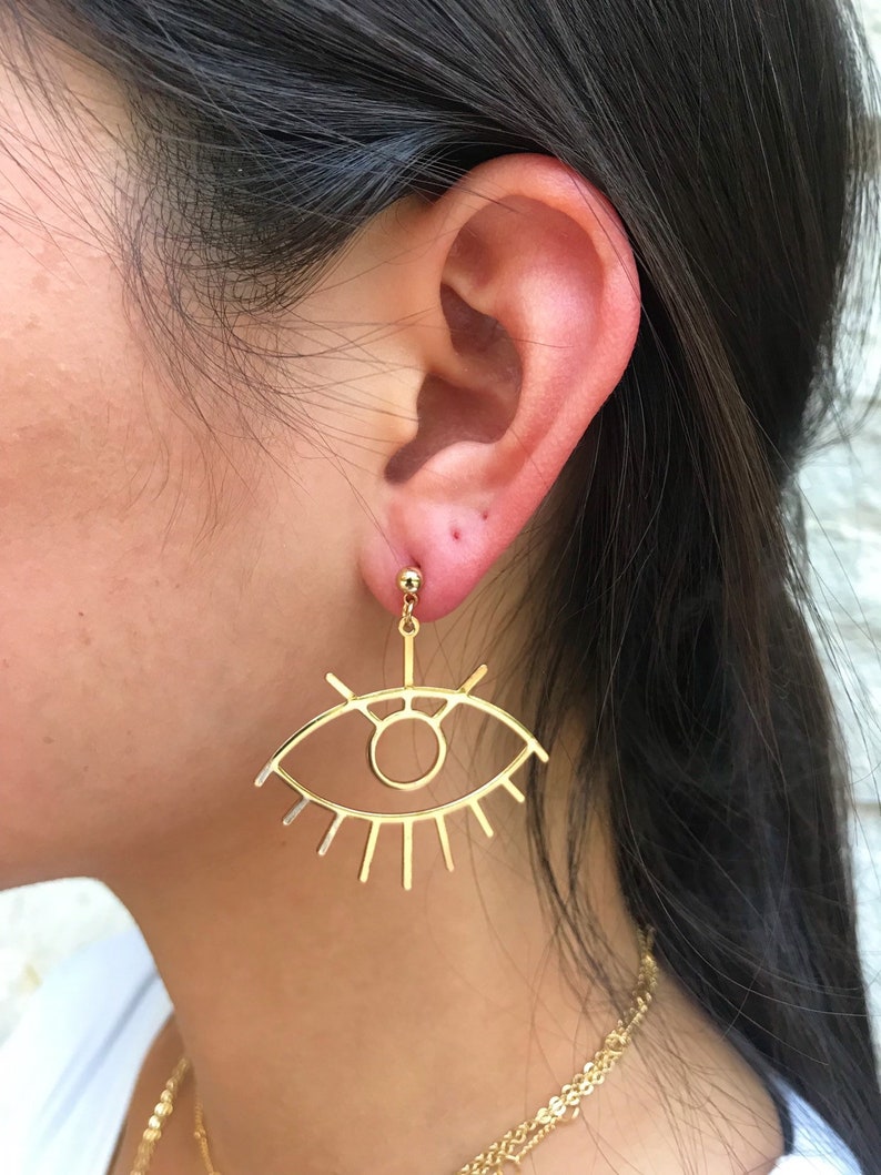 May include: Gold eye-shaped earrings with a delicate outline and radiating lines. The earrings are dangling from the earlobe.