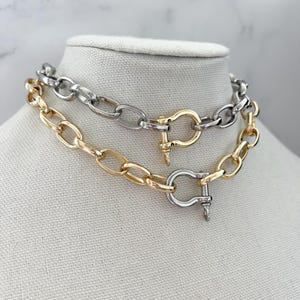 Puede incluir: Dos collares de cadena exhibidos en un maniqu&iacute;. Uno es dorado, el otro plateado. Ambos presentan eslabones ovalados grandes y un cierre de broche. El collar dorado est&aacute; debajo del collar plateado.
