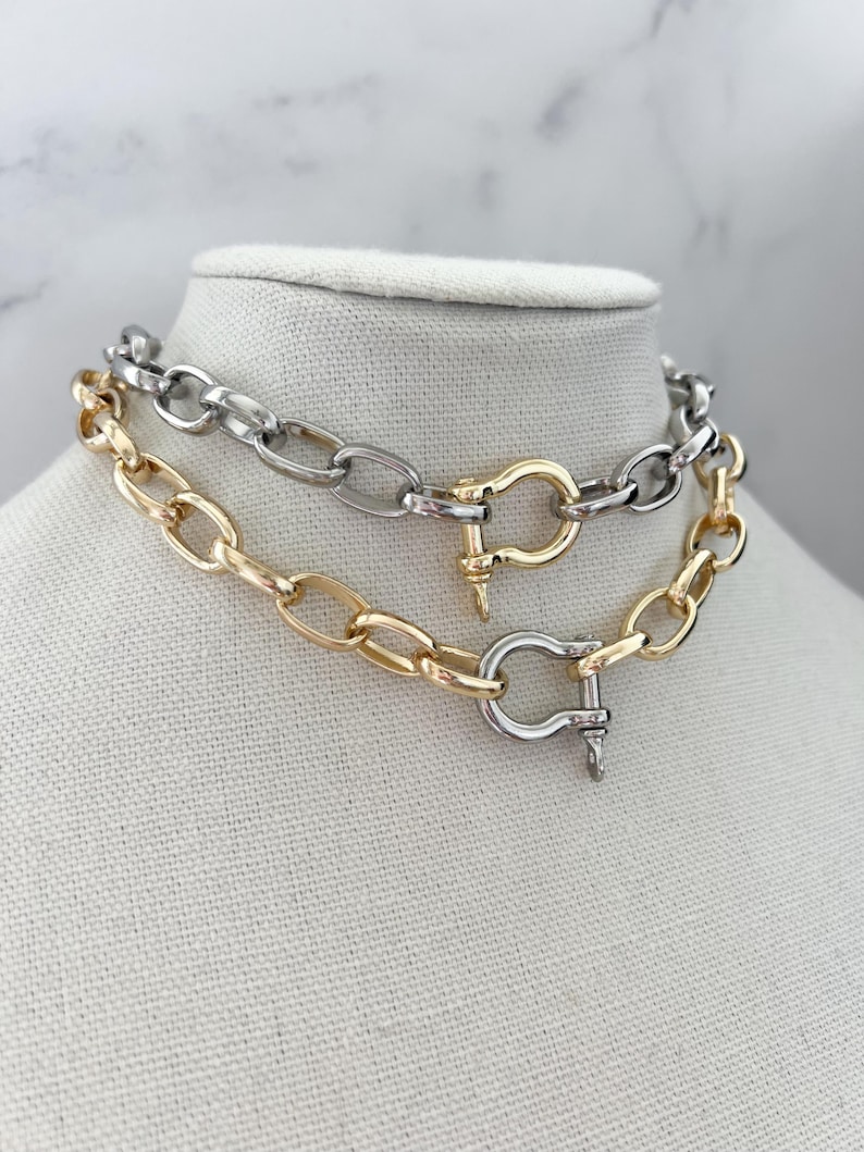 Puede incluir: Dos collares de cadena exhibidos en un maniqu&iacute;. Uno es dorado, el otro plateado. Ambos presentan eslabones ovalados grandes y un cierre de broche. El collar dorado est&aacute; debajo del collar plateado.