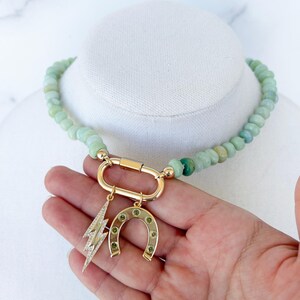 May include: A green gemstone beaded necklace with a gold clasp and charms. The charms include a horseshoe and a lightning bolt.