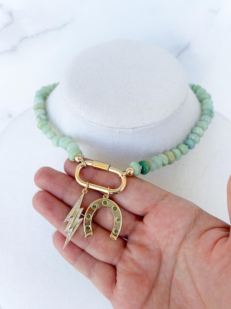 May include: A green gemstone beaded necklace with a gold clasp and charms. The charms include a horseshoe and a lightning bolt.