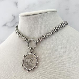 May include: A silver-toned necklace featuring a thick, textured chain connected to a circular pendant. The pendant displays a relief of a horse in the centre, surrounded by small, evenly spaced metallic spheres along the outer edge of the frame.