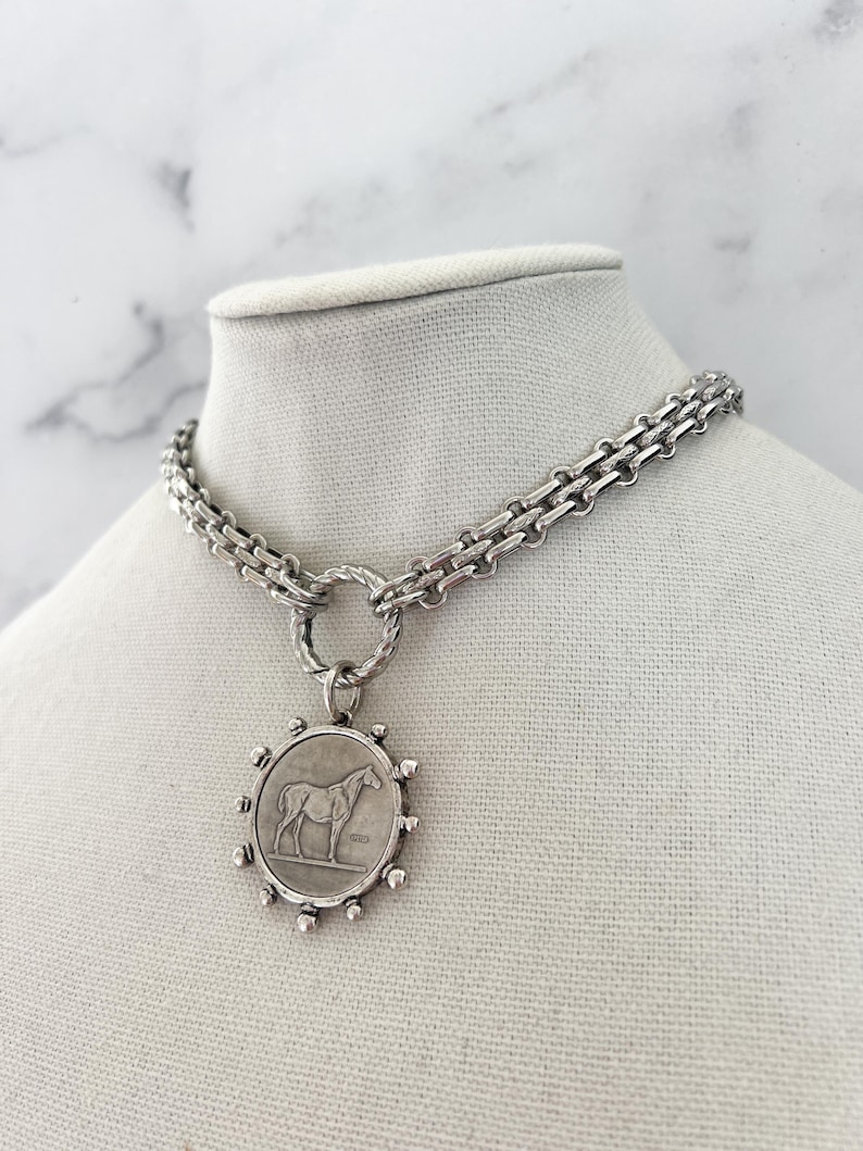 May include: A silver-toned necklace featuring a thick, textured chain connected to a circular pendant. The pendant displays a relief of a horse in the centre, surrounded by small, evenly spaced metallic spheres along the outer edge of the frame.