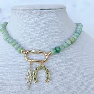 May include: A green jade stone beaded necklace with a gold charm featuring a horseshoe and lightning bolt. The charms are accented with small green stones.