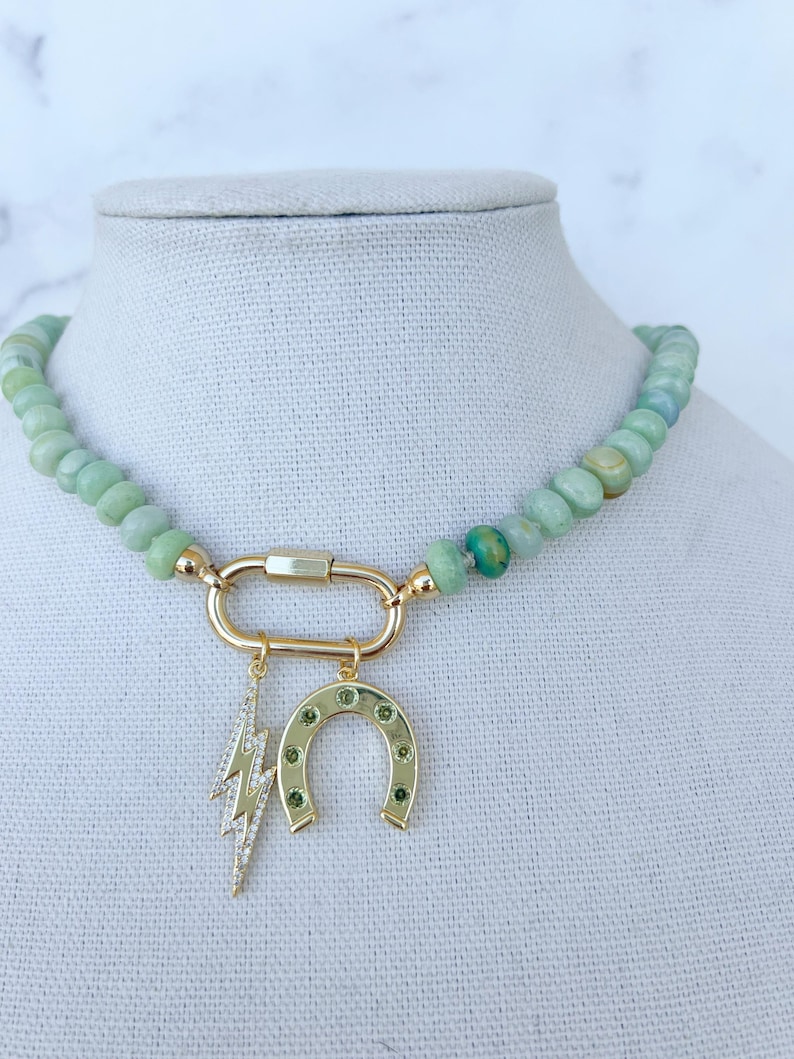 May include: A green jade stone beaded necklace with a gold charm featuring a horseshoe and lightning bolt. The charms are accented with small green stones.