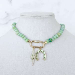 May include: A green jade beaded necklace with a gold charm featuring a horseshoe and lightning bolt. The necklace is on a white mannequin.