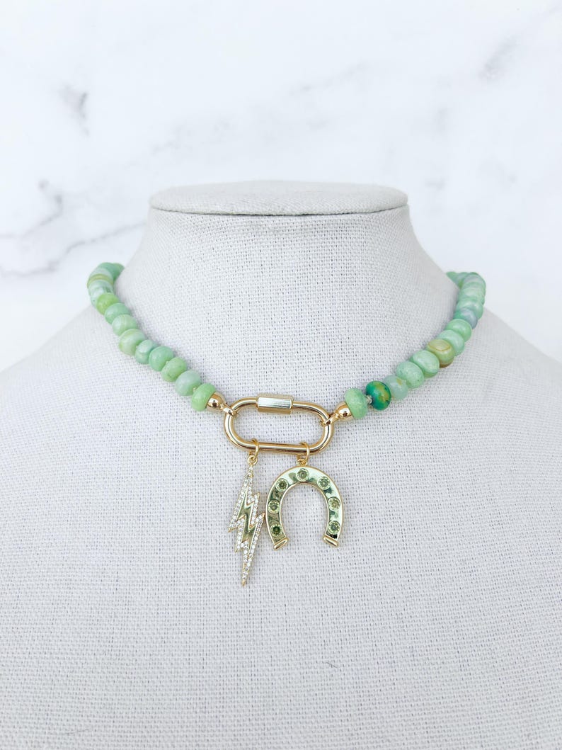May include: A green jade beaded necklace with a gold charm featuring a horseshoe and lightning bolt. The necklace is on a white mannequin.