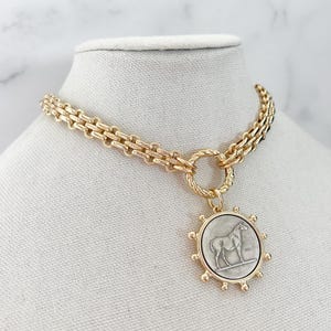 May include: A gold-toned chain necklace featuring a circular pendant with a silver-coloured horse relief. The pendant is framed by small gold spheres and attached to the chain by a twisted gold ring. The chain consists of interlocking rectangular links.