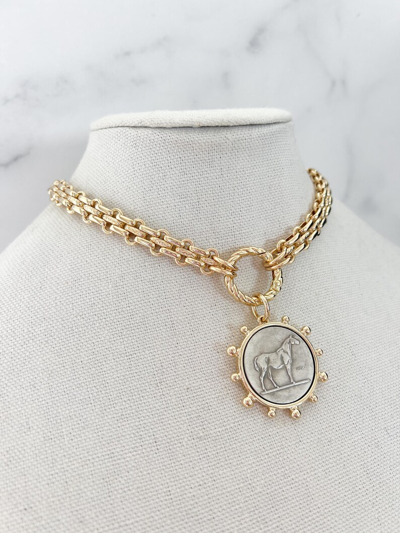 May include: A gold-toned chain necklace featuring a circular pendant with a silver-coloured horse relief. The pendant is framed by small gold spheres and attached to the chain by a twisted gold ring. The chain consists of interlocking rectangular links.