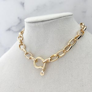 Puede incluir: Collar de cadena dorado con eslabones ovalados grandes y un cierre en forma de coraz&oacute;n. El collar se exhibe sobre un busto de maniqu&iacute; blanco sobre un fondo de m&aacute;rmol. Una pieza audaz.