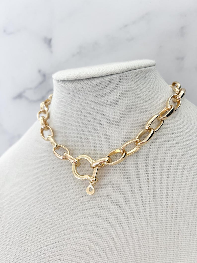 Puede incluir: Collar de cadena dorado con eslabones ovalados grandes y un cierre en forma de coraz&oacute;n. El collar se exhibe sobre un busto de maniqu&iacute; blanco sobre un fondo de m&aacute;rmol. Una pieza audaz.
