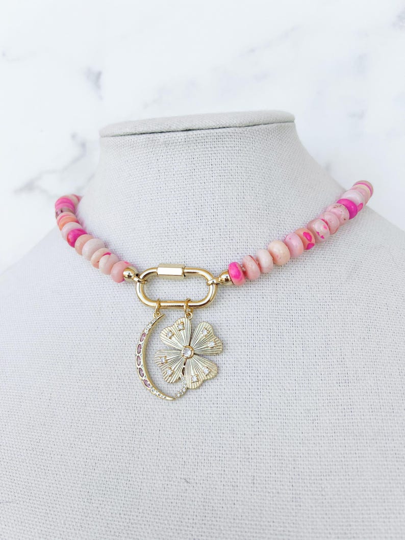 May include: A pink and white beaded necklace with a gold clasp and a charm featuring a crescent moon and a flower with rhinestones.
