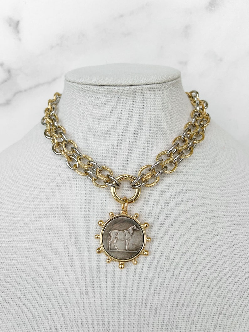 May include: A gold and silver chain necklace with a round pendant featuring a horse design. The pendant has a gold rim with small gold balls around the edge.