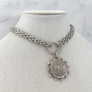May include: A silver-toned necklace featuring a thick, textured chain connected to a circular pendant. The pendant displays a relief of a horse in the centre, surrounded by small, evenly spaced silver spheres along the outer edge of the frame.