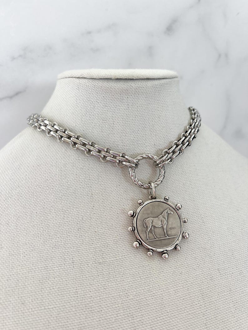 May include: A silver-toned necklace featuring a thick, textured chain connected to a circular pendant. The pendant displays a relief of a horse in the centre, surrounded by small, evenly spaced silver spheres along the outer edge of the frame.