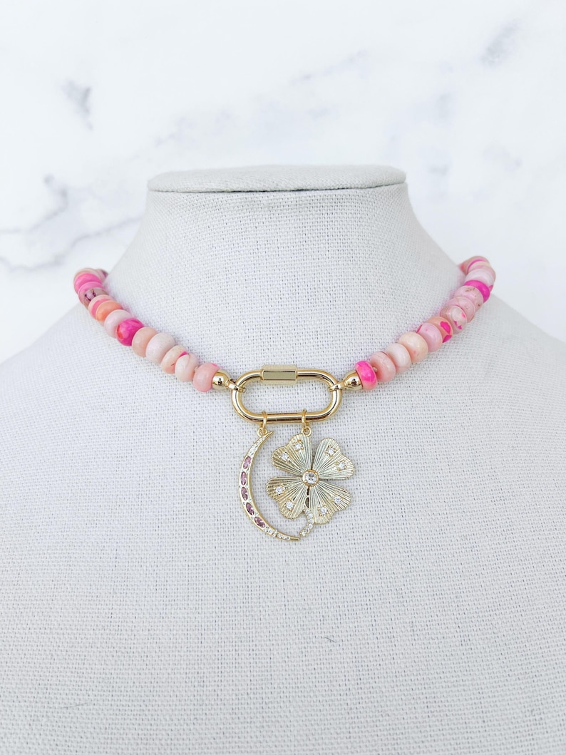 May include: A pink and white beaded necklace with a gold clasp and a charm featuring a crescent moon and a flower with rhinestones.